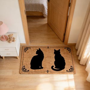 Könnte beinhalten: Eine rechteckige Fußmatte aus Naturfasern mit zwei schwarzen Katzen-Silhouetten. Die Matte hat einen dekorativen Rand mit Schnörkeln und liegt in einem Türrahmen auf einem Holzboden.