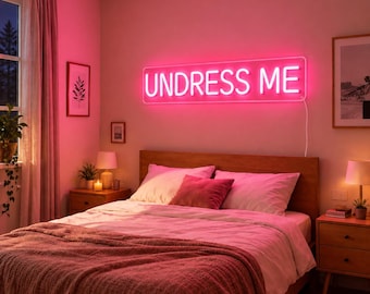 May include: A rectangular neon sign mounted on a wall above a bed, displaying the text UNDRESS ME in bright pink illuminated letters. The sign emits a vibrant pink glow that casts light across the bedroom, highlighting the bedding and decor.