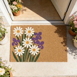 May include: A rectangular doormat made of natural fibers. It features a floral design with white daisies, purple irises, and green stems. The mat is placed in front of a white door, with potted plants on either side.