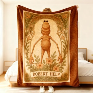 May include: A brown plush blanket with a quirky illustration of a creature with long arms and the text "ROBERT, HELP". The design is framed by floral and art nouveau-inspired elements, with a light beige background and a dark brown border.