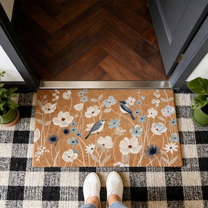 Può includere: Un zerbino rettangolare con un design floreale e di uccelli. Il tappeto presenta fiori bianchi e azzurri con due uccelli blu su uno sfondo marrone. Il tappeto è posizionato su un tappeto a scacchi in bianco e nero.