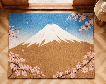 May include: A rectangular doormat featuring a textured, tufted design of Mount Fuji with a snow-capped peak, a brown mountain base, and a bright blue sky. Pink cherry blossom branches frame the top right and bottom left corners of the mat.