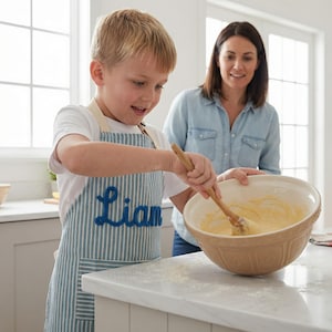Puede incluir: Un ni&ntilde;o con un delantal a rayas azules y blancas con el nombre "Liam" bordado, mezclando masa en un cuenco beige grande con una cuchara de madera. Una mujer est&aacute; cerca, sonriendo. La escena tiene lugar en una cocina luminosa.