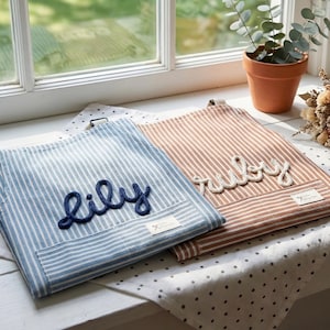 Puede incluir: Dos delantales a rayas personalizados, uno azul y blanco, el otro marr&oacute;n y blanco. El delantal azul tiene "LILY" bordado en azul marino, y el delantal marr&oacute;n tiene "ruby" bordado en blanco. Ambos tienen un peque&ntilde;o bolsillo.