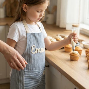 Puede incluir: Un delantal a rayas azules y blancas con el nombre "Emma" bordado en la parte delantera. Una persona est&aacute; decorando cupcakes con glaseado. El delantal tiene un bolsillo y correas beige. Los cupcakes est&aacute;n en una encimera de madera.