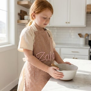 Puede incluir: Un delantal a rayas marrones y blancas con el nombre "Zaha" bordado en rosa. El delantal tiene un bolsillo delantero y se usa mientras se sostiene un cuenco y un batidor blancos. El delantal est&aacute; dise&ntilde;ado para uso en la cocina.