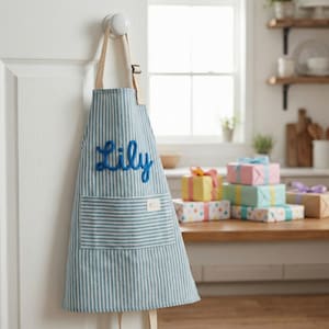 Puede incluir: Delantal a rayas azules y blancas con el nombre "Lily" bordado en azul. El delantal tiene un bolsillo y est&aacute; colgado de una puerta blanca. Varios regalos envueltos est&aacute;n sobre una mesa de madera en el fondo.