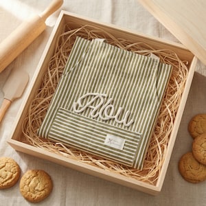 Puede incluir: Una caja de regalo de madera contiene un delantal a rayas verdes y blancas con el nombre "Abou" bordado. Tambi&eacute;n hay un rodillo, una esp&aacute;tula y galletas. El delantal tiene un bolsillo con una peque&ntilde;a etiqueta.