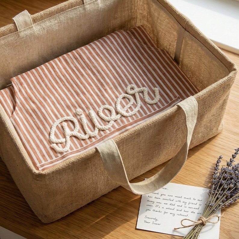 Puede incluir: Una bolsa de mano de yute beige con asas, que contiene una tela a rayas marr&oacute;n y blanca. La palabra "River" est&aacute; escrita con cuerda blanca. Se ve una tarjeta peque&ntilde;a y ramitas de lavanda.