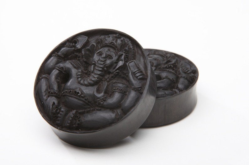 Pode incluir: Dois medidores de orelha de madeira preta com uma imagem esculpida da divindade hindu Ganesha. Os medidores s&atilde;o redondos e t&ecirc;m um acabamento liso e polido.