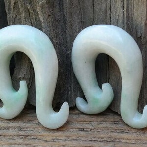 Puede incluir: Un par de dilatadores de oreja de jade verde en forma de gancho. Los dilatadores están hechos de jade liso y pulido y tienen un color verde claro.