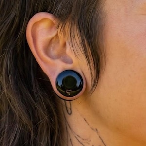 May include: A smooth, circular black ear plug gauge inserted into a stretched earlobe. The jewelry features a polished, reflective surface with a solid black color, creating a simple and uniform appearance within the ear piercing.