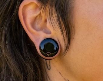 May include: A smooth, circular black ear plug gauge inserted into a stretched earlobe. The jewelry features a polished, reflective surface with a solid black color, creating a simple and uniform appearance within the ear piercing.