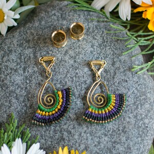 May include: A pair of gold-toned earrings with a spiral design and a fan-shaped beaded fringe in shades of green, purple, and black. The earrings are hanging from a gold chain with a clasp closure. Two gold-toned ear gauges are also included.