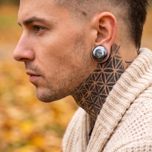 May include: A smooth, polished, dark metallic circular ear plug worn in a stretched earlobe. The jewelry features a reflective, gunmetal-colored surface with a slightly domed profile, securely fitted within the ear piercing.