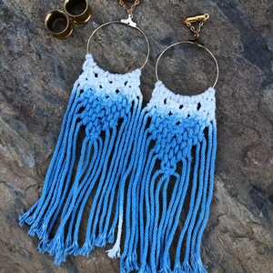 Op de afbeelding: Een paar blauwe en witte macramé oorbellen met gouden hoepels en kettingverbinders. De oorbellen zijn gemaakt met een gradiënt van blauw en wit garen.
