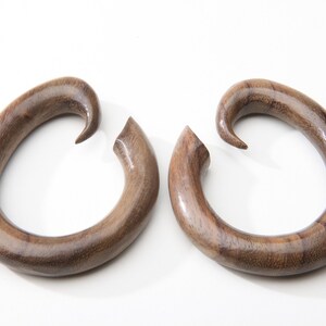 May include: Two dark brown wooden earrings, each with a curved, teardrop shape. The earrings have a smooth, polished finish and a hook-like design at the top. They are presented against a white backdrop.