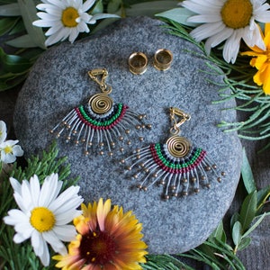 May include: A pair of beaded earrings with a spiral design and a gray, red, and green color scheme. The earrings are on a gray stone with two gold metal plugs.