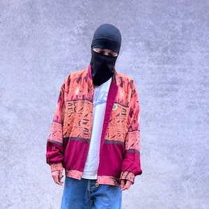 Puede incluir: Chaqueta bomber estampada con un panel inferior burdeos, combinada con vaqueros azules y zapatos marrones. La chaqueta presenta un diseño naranja y rojo con símbolos de tipo jeroglífico. Se ve una camiseta blanca con el logo de Nike.