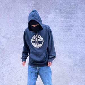 May include: Navy blue hoodie with a white and tan tree logo, paired with light blue jeans and brown boots. The hoodie has a front pocket and a drawstring hood. The jeans are a classic fit.