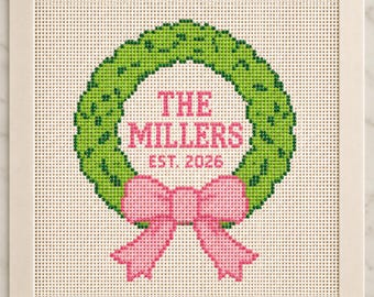 May include: A square needlepoint canvas featuring a green wreath design with a pink bow at the bottom. The center of the wreath contains the pink text THE MILLERS EST. 2026. The canvas is mounted on a white frame with taped edges.