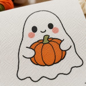 May include: A cross-stitch pattern on white Aida fabric featuring a white ghost with black eyes, a small smile, and pink cheeks. The ghost is holding a bright orange pumpkin with a green stem, outlined in black thread throughout the design.