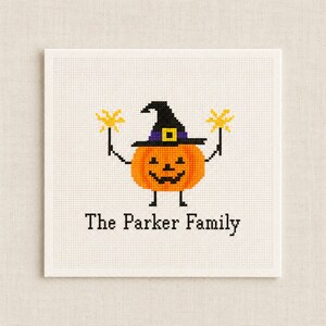 Peut inclure: Un motif de point de croix sur tissu blanc représentant une citrouille orange portant un chapeau de sorcière noir avec une bande violette et une boucle jaune. La citrouille tient deux cierges magiques jaunes et le texte "The Parker Family" est brodé en dessous.
