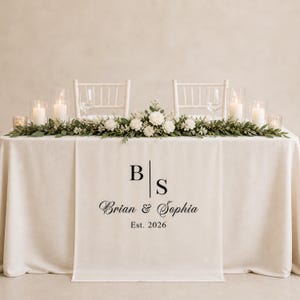 Könnte beinhalten: Eine Hochzeitstafel mit weißer Tischdecke, Blumenschmuck und Kerzen. Ein Tischläufer zeigt die Initialen "B S", die Namen "Brian & Sophia" und das Jahr "Est. 2026". Im Hintergrund stehen zwei weiße Stühle.