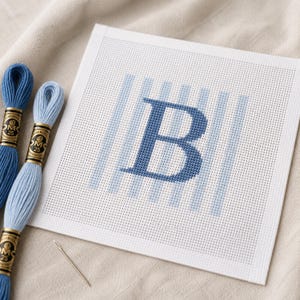 Pode incluir: Uma tela quadrada branca de ponto de agulha com uma letra B azul-escura centrada sobre listras verticais azul-claras. Dois novelos de linha de bordar azul com etiquetas douradas e uma agulha de costura prateada estão à esquerda.
