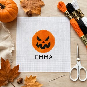 Könnte beinhalten: Ein quadratischer weißer Kreuzstichstoff mit einem zentrierten orangefarbenen Kürbisgesicht mit schwarzen Augen und einem gezackten Mund, darunter der Name EMMA in Schwarz gestickt. Das Design ist umgeben von einem Kürbis, Herbstblättern, Stickgarn und einer Schere.
