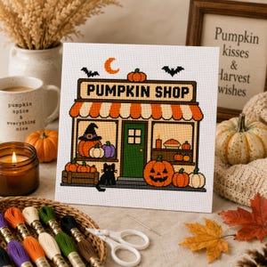 Peut inclure: Un motif de point de croix sur tissu blanc représentant une devanture de magasin marquée PUMPKIN SHOP avec un auvent orange, une porte verte, un chat noir et diverses citrouilles. Le motif inclut un croissant de lune et deux chauves-souris.
