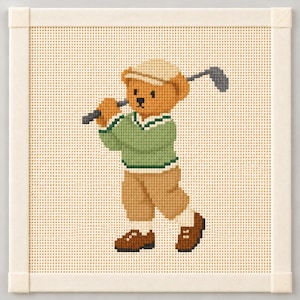 May include: A cross-stitch embroidery piece featuring a light brown teddy bear wearing a beige cap, a green sweater with white trim, and tan trousers. The bear is holding a grey golf club over its shoulder, set against a cream-colored fabric background.