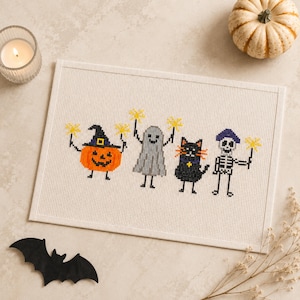 May include: A rectangular cross-stitch embroidery on white fabric featuring four characters: an orange pumpkin with a black hat, a gray ghost, a black cat, and a white skeleton. Each character holds a yellow star-shaped sparkler.
