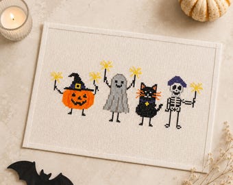 May include: A rectangular cross-stitch embroidery on white fabric featuring four characters: an orange pumpkin with a black hat, a grey ghost, a black cat, and a white skeleton. Each character holds a yellow star-shaped sparkler.