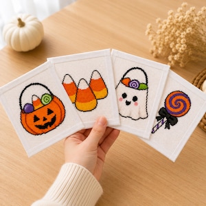 May include: Four square white fabric panels featuring cross-stitch designs of Halloween motifs: a jack-o'-lantern bucket filled with sweets, three pieces of orange, yellow, and white candy corn, a white ghost holding a bucket, and a purple and orange lollipop.
