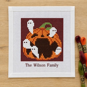 Peut inclure: Une broderie au point de croix carrée sur tissu blanc représentant une citrouille orange avec un intérieur marron foncé et quatre petits fantômes blancs. Le texte "The Wilson Family" est brodé en marron foncé sous le motif.