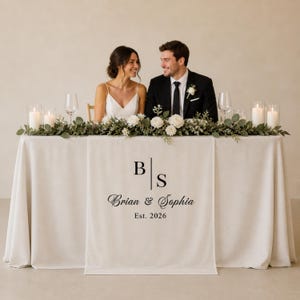 May include: A white table runner with the initials "B" and "S" and the names "Brian & Sophia" and the year "Est. 2026" printed in black. The table is set for a wedding with a floral arrangement and candles.