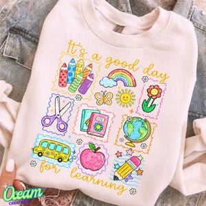 Peut inclure: Un sweat-shirt rose clair avec une impression graphique colorée portant le texte "It's a good day for learning". Le motif comprend des illustrations de crayons, un arc-en-ciel, un papillon, un soleil, une fleur, des ciseaux, des livres, un globe, un bus, une pomme et un crayon.