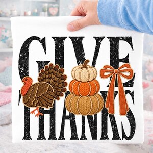 Op de afbeelding: Een wit t-shirt met de zwarte, verweerde tekst GIVE THANKS. Het ontwerp bevat een bruine kalkoen, een stapel van drie pompoenen in beige en oranje tinten, en een oranje strik, allemaal weergegeven met een textuur in borduurstijl.
