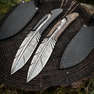 May include: Two ornate knives with patterned blades and wooden handles rest on a tree stump. The blades have a unique design with etched lines. Black leather sheaths with textured patterns are also visible.