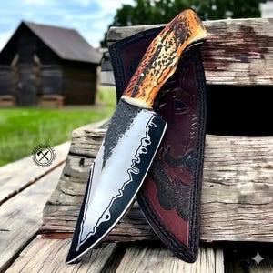 May include: A handcrafted knife with a textured blade and an antler handle. The knife is displayed with a leather sheath, resting on a wooden surface. The blade has a unique design along the edge. The handle is brown and tan.