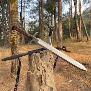 May include: A large silver sword with a brown handle and a smaller knife are displayed on a tree stump. The sword has a long, straight blade and a crossguard. A black sheath and leather strap are also visible.