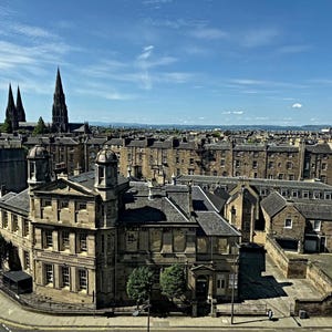 May include: An aerial view of a city with stone buildings under a bright blue sky. The image features a large building with a domed roof and a church with tall spires in the background. The architecture is classic.