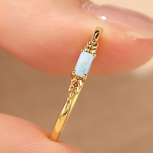May include: A thin gold ring featuring a rectangular, light blue opal stone set horizontally. The metal band displays intricate, textured leaf-like detailing on both sides of the central stone, creating a delicate and refined appearance.