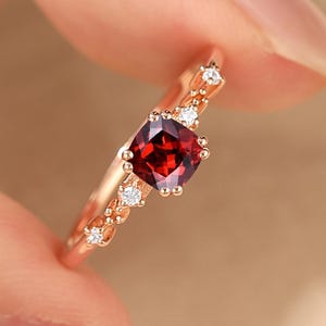 May include: Rose gold ring featuring a square, deep red gemstone as the centerpiece. The band is adorned with small, clear accent stones and delicate filigree details. The ring is held up by a hand, showcasing its design and craftsmanship.