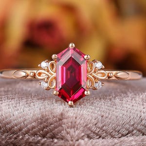 May include: Rose gold ring featuring a vibrant, faceted, hexagonal ruby gemstone. The ring has delicate filigree detailing with small, sparkling accent stones on either side of the ruby. The band is slender and polished, showcasing the central stone.