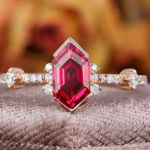 May include: A gold ring featuring a large, faceted, red gemstone in a unique geometric shape. The ring is adorned with small, clear accent stones along the band and around the central stone, creating a sparkling effect. The ring is set on a velvet surface.