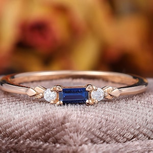 May include: A rose gold ring featuring a central baguette-cut blue sapphire flanked by two small round-cut white diamonds. The band has a delicate, slender profile with decorative metal leaf-like accents on either side of the gemstone setting.