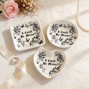 May include: Three white ceramic trinket dishes in square, round, and heart shapes. Each dish features a black skull and butterfly design with the text "I Could Be Meaner". The heart-shaped dish has a gold rim.