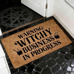 May include: A rectangular doormat with the text "WARNING: WITCHY BUSINESS IN PROGRESS" in black. The mat is brown and features two cauldron illustrations. The doormat is placed in front of a white door on a black tiled floor.